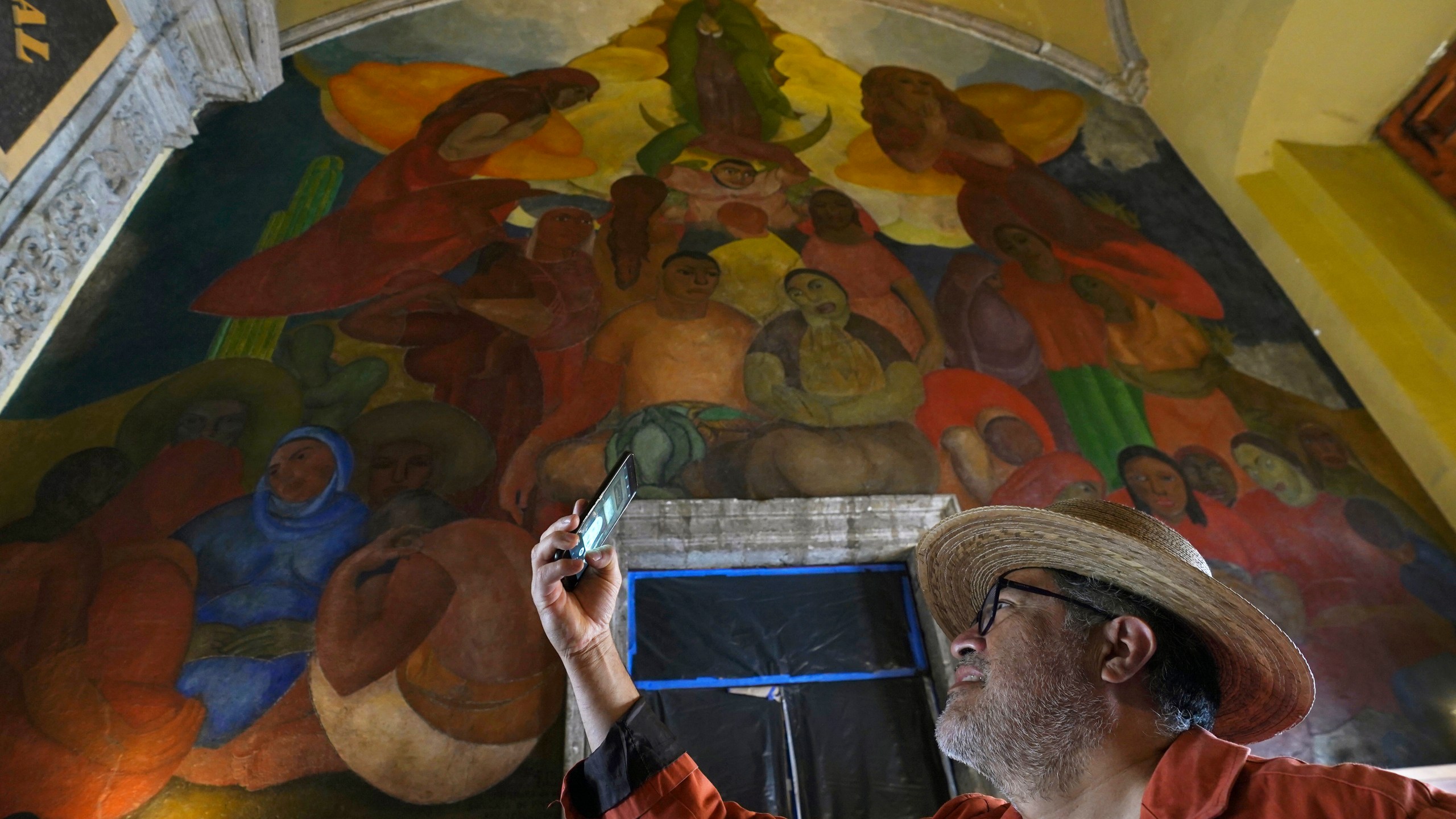 A tourist takes a photo backdropped by the "Alegoria de la Virgen de Guadalupe" mural, in the main entrance of the Antiguo Colegio de San Ildefonso, in Mexico City, Wednesday, April 26, 2023. The mural was created by Mexican artist Fermin Revueltas between 1922 and 1923, when the walls of San Ildefonso became the canvases where the muralist movement came to life. (AP Photo/Marco Ugarte)